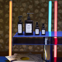 Thumbnail for Set of black skincare bottles with labels on a blue shelf against a floral-patterned wall.