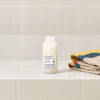 Thumbnail for White bottle with a label on a tiled bathroom shelf next to folded towels.