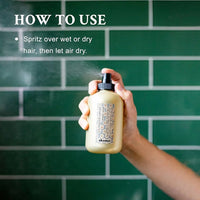 Thumbnail for Hand holding a hair product spray bottle with text on how to use it against a green tiled wall.