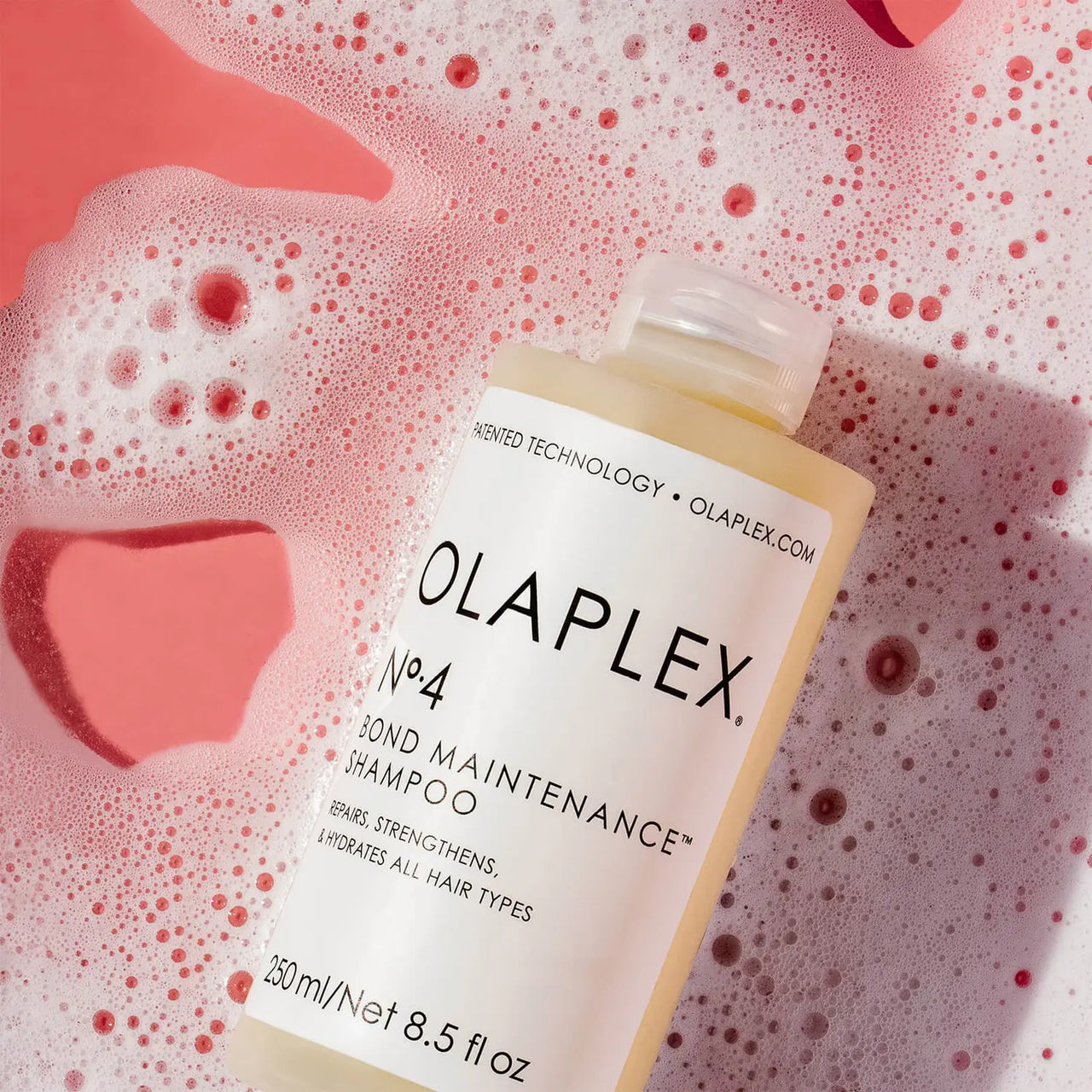 A bottle of Olaplex No. 4 Bond Maintenance Shampoo placed on pink and white foamy water with a pink heart-shaped soap dish, showing the product label clearly.
