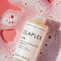 Thumbnail for A bottle of Olaplex No. 4 Bond Maintenance Shampoo placed on pink and white foamy water with a pink heart-shaped soap dish, showing the product label clearly.
