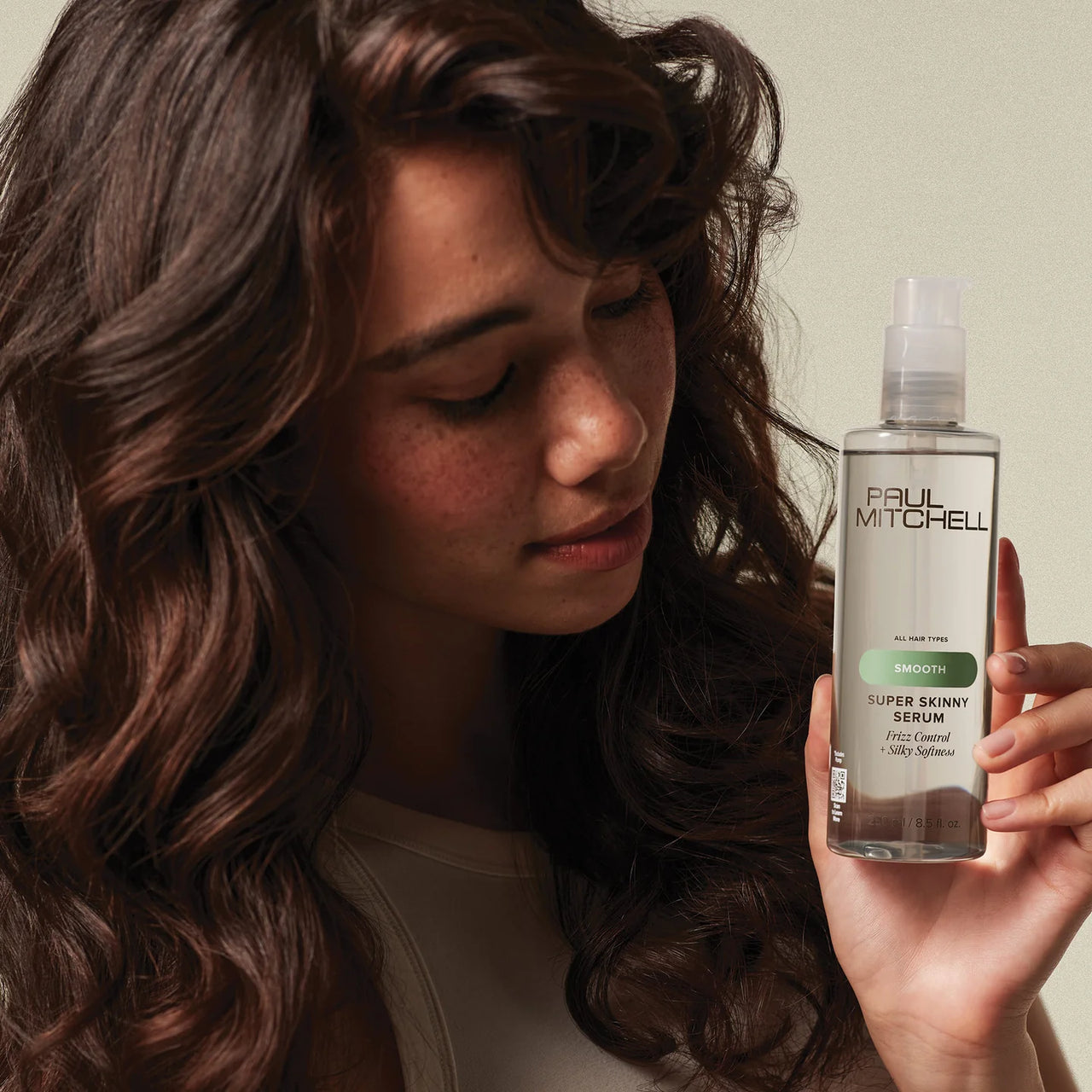 Woman holding a bottle of Paul Mitchell Super Skinny Serum with a neutral background