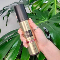 Thumbnail for A hand holding a gold and black ghd body paint bottle, set against a background of large green monstera leaves.