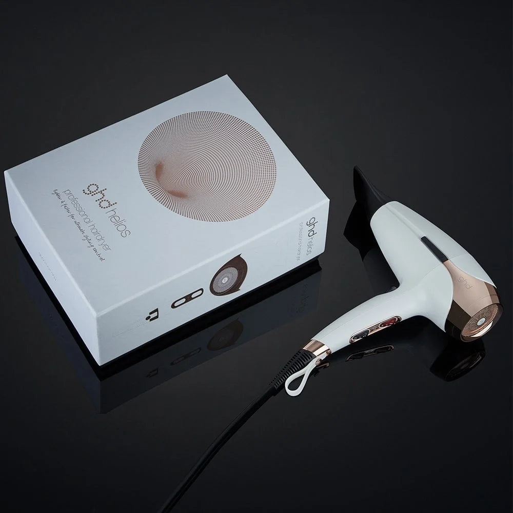 A white and rose gold hair dryer shown next to its white retail box on a glossy black reflective surface. The box features the brand name 'ghd' and 'Hair Pro'.