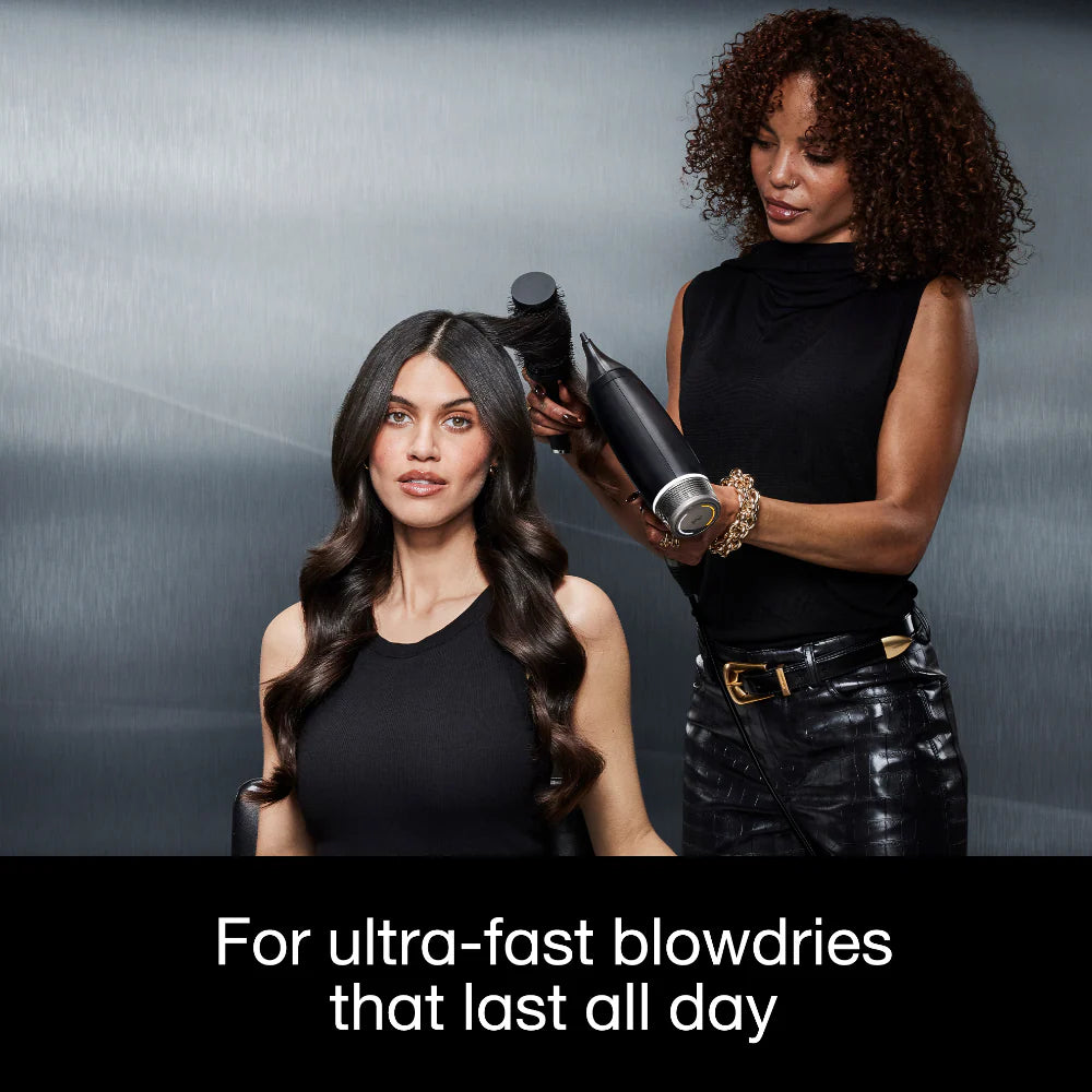 Woman getting her hair styled by another woman with a hair dryer against a gray background.