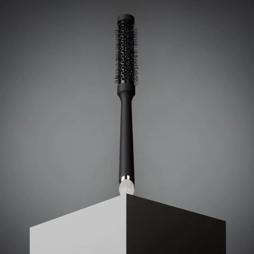 A black round hairbrush with black bristles and a silver-toned metal neck accent, standing upright on a geometric display stand against a dark gray background.