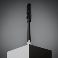 Thumbnail for A black round hairbrush with black bristles and a silver-toned metal neck accent, standing upright on a geometric display stand against a dark gray background.