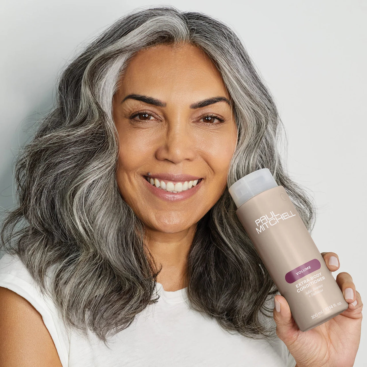 Woman with gray hair holding a bottle of Paul Mitchell product against a plain background